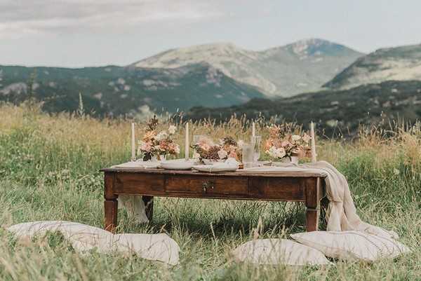 low dark wood coffee table with cream fabric, candles and pastel floral arrangement in field with mountain views