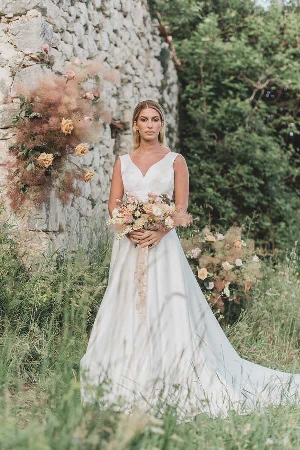 bride stands in front of stone castle ruins holding cream bouquet