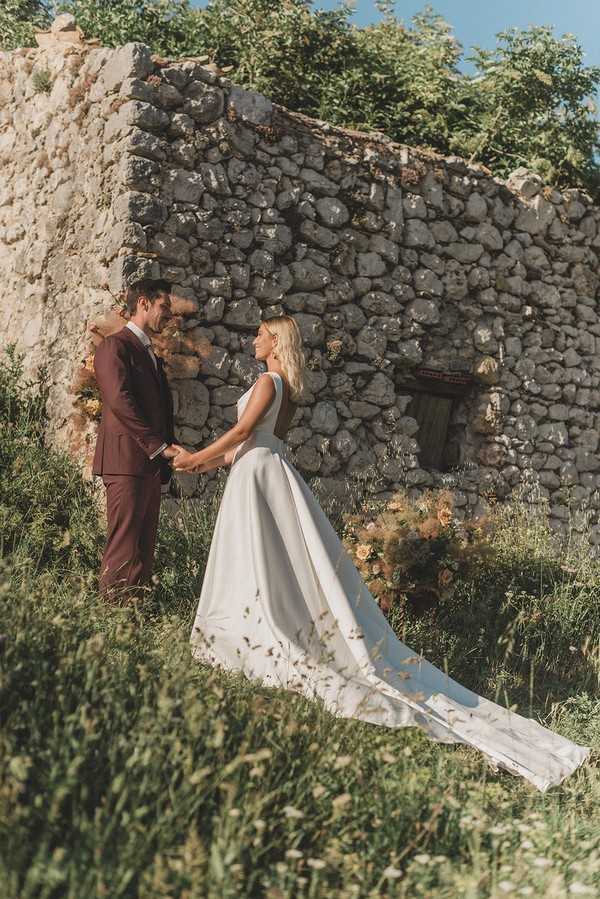 bride and groom meet in front of stone ruins her dress trails behind her and he wears a burgundy suit