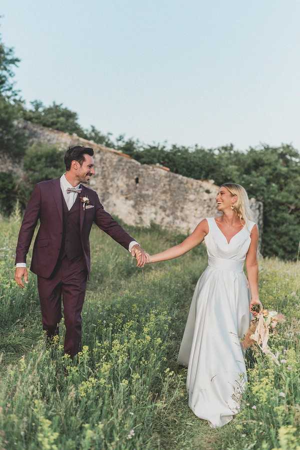 bride and groom hold hands and walk through green field with stone ruins visible behind them