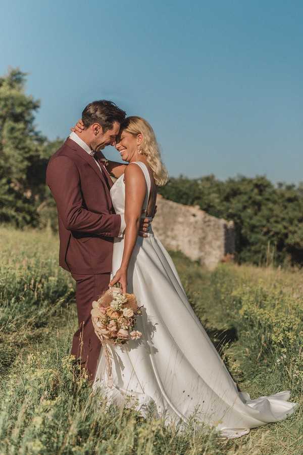 bride and groom hug and smile standing in a green field with stone ruins behind them 