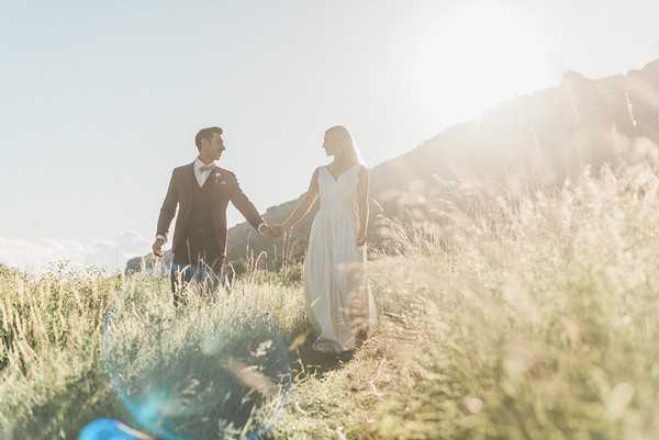 bride and groom hold hands and walk through sundrenched yellowing field