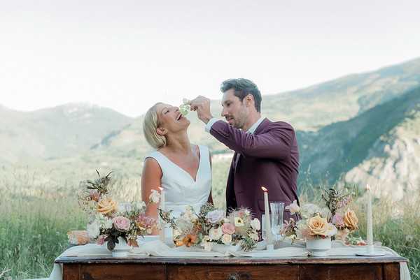 groom feeds his bride grapes as they sit on the ground in front of small decorated wedding table with rolling mountain views behind them