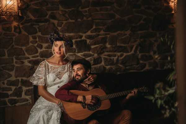 bride sits with groom on his guitar in front of stone wall