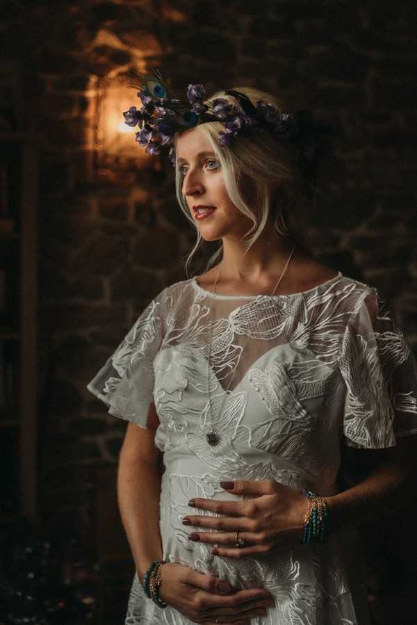 pregnant bride in lace dress and peacock feather crown holds her belly