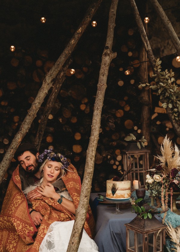 wooden log teepee frames bride and groom hugging under red and gold quilt