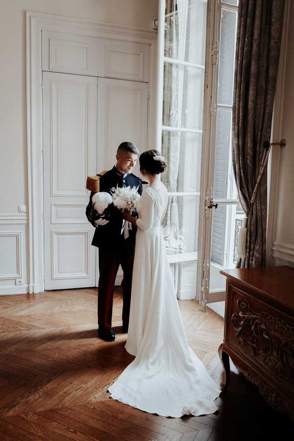 bride in retro low back wedding dress meets her groom in full military uniform at the window of a french chateau