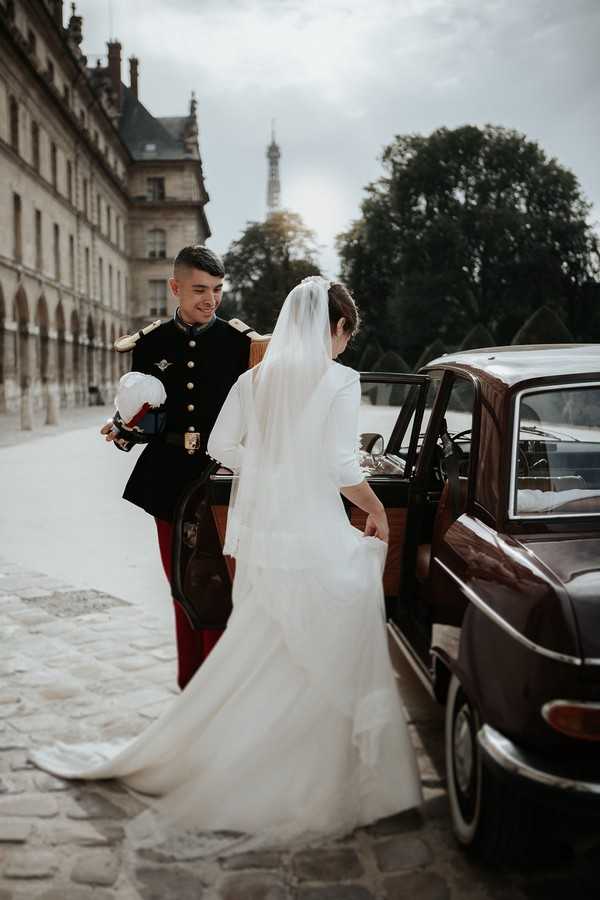 Groom holds door to red vintage car open for his bride