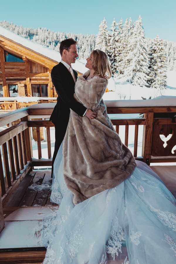 bride wrapped in a fur blanket and groom in black suit with snow and trees behind them