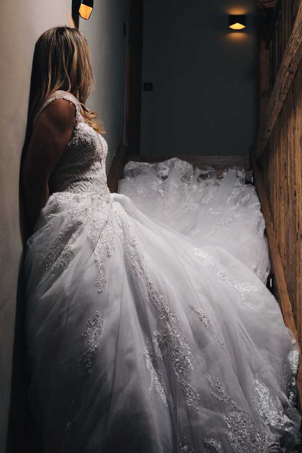 bride stands in a shadowy stair well and her dress trails up the stairs behind her, she is turned away from the camera