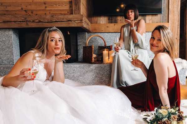 bride blows a kiss to the camera holding a glass of champagne with her bridesmaids who are wearing red and pale green