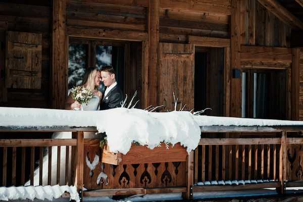 bride and groom smile on the wooden balcony of a snow covered private chalet in the French Alps