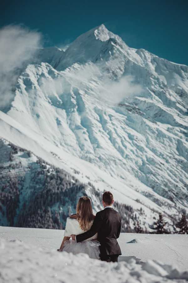 large snow covered mountain makes up the majority of this photo of a bride and groom arm and arm in the snow