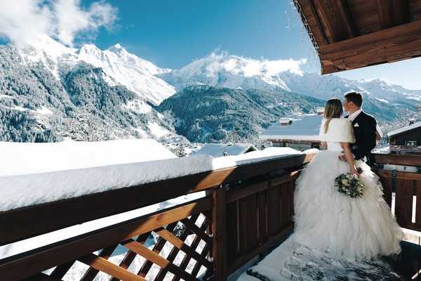 bride wears white fur wrap and groom wears black suit on the snowy balcony of a French chalet overlooking snow covered mountains