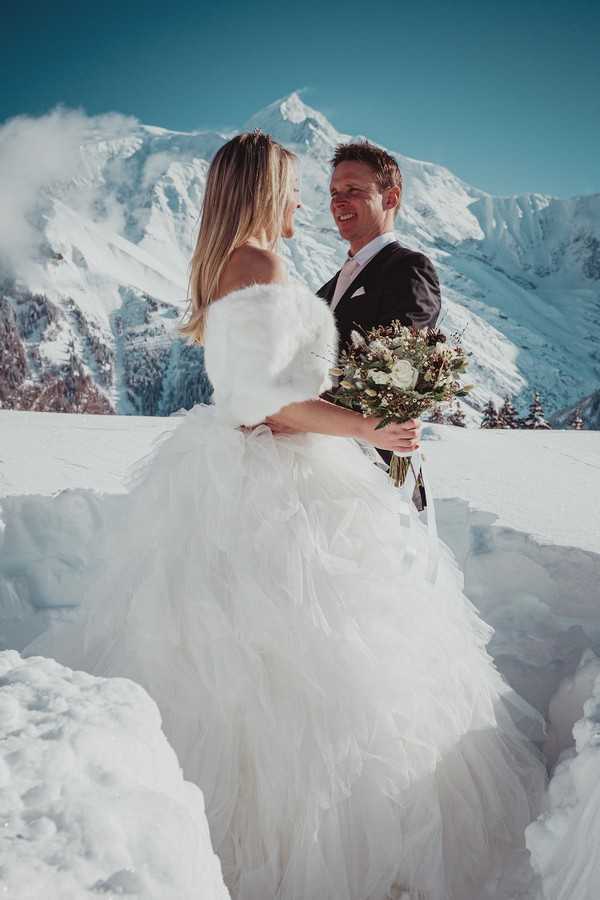 bride in white fur wrap smiles at her groom with snowy mountain in the background. they are standing waist high in white snow