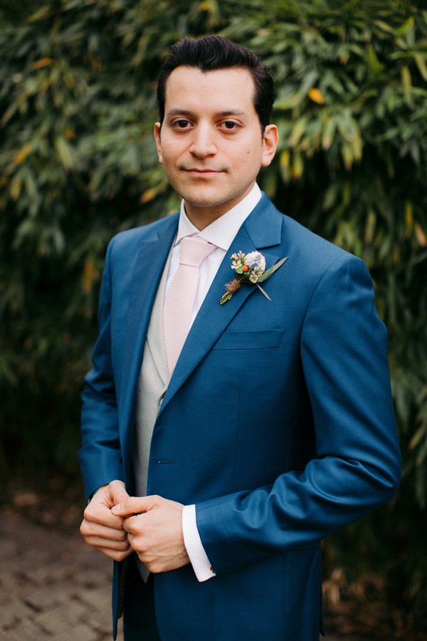 Groom in a blue suit with floral buttonhole in front of shrub