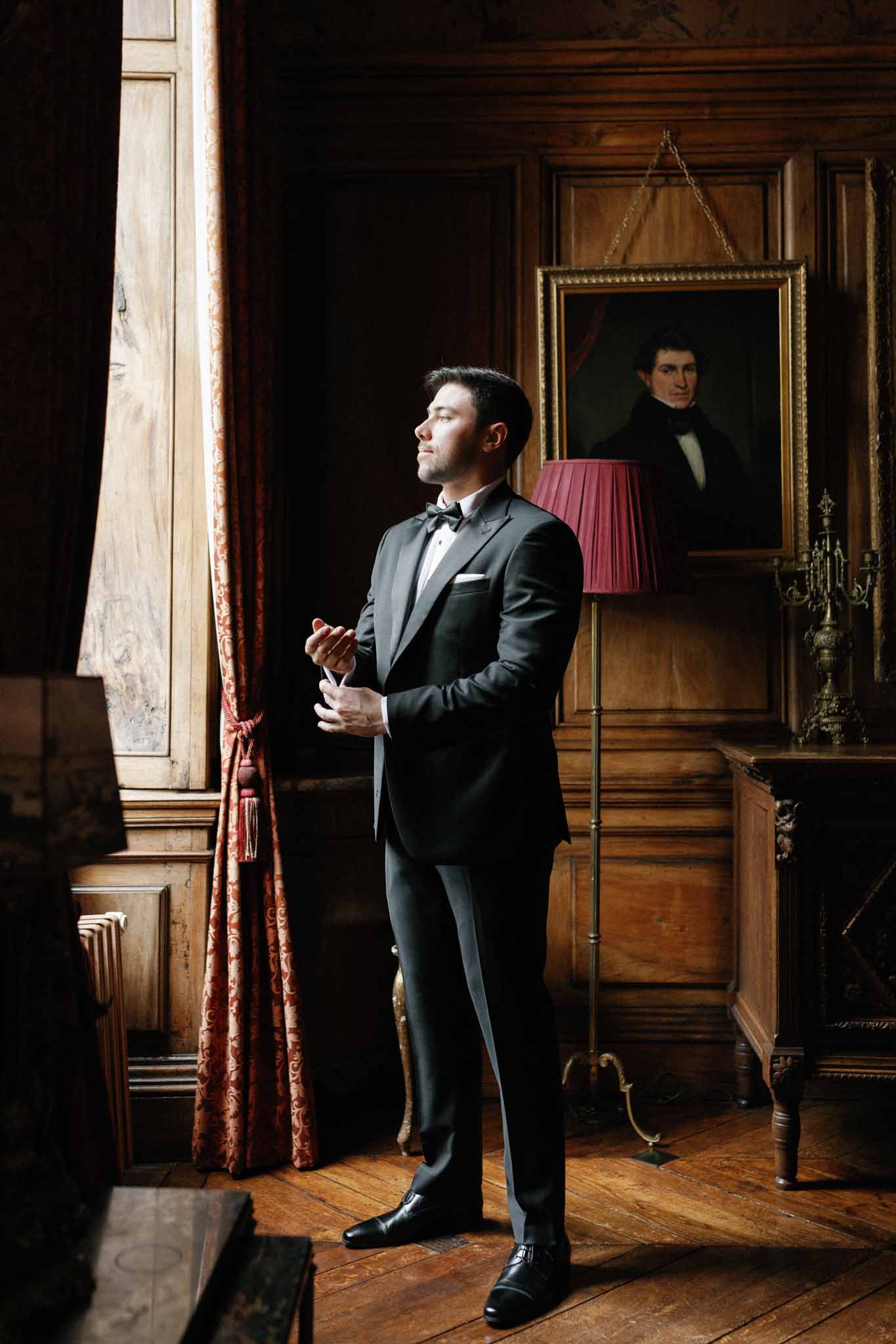 Groom in charcoal tuxedo standing in elegant wood-paneled room with period furnishings
