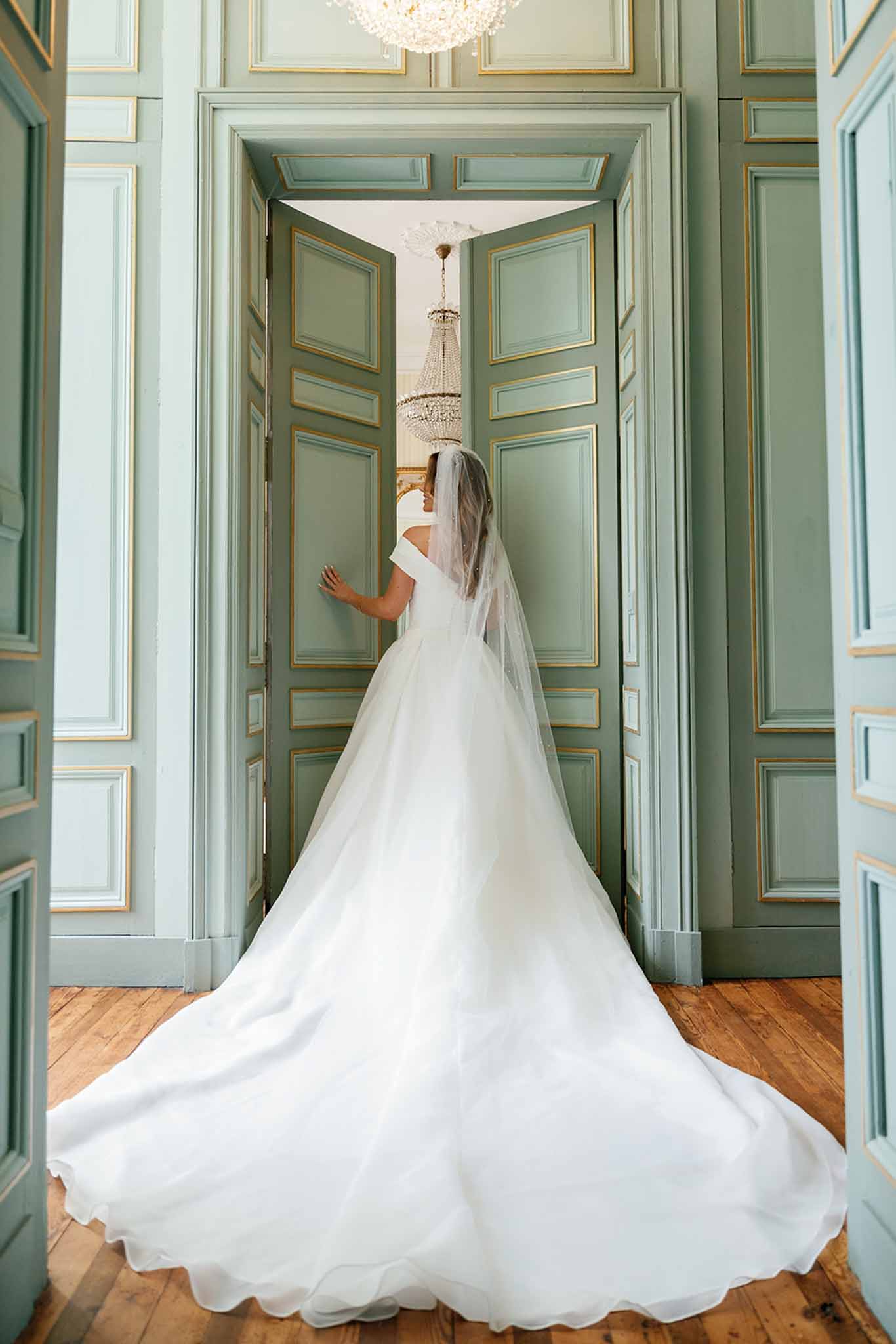 Bride in ivory gown opening sage green doors in classical interior with chandelier