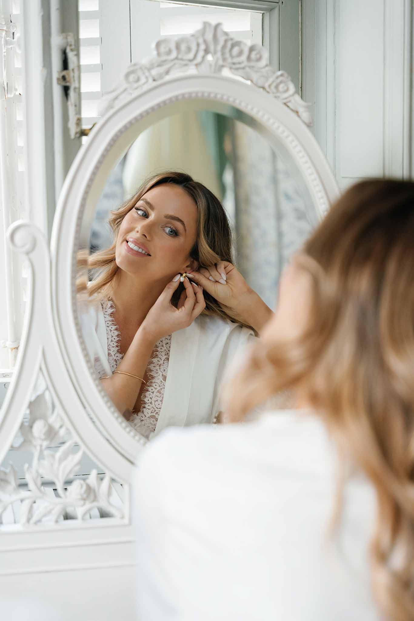 Bride adjusting earrings in ornate mirror during wedding preparation indoors