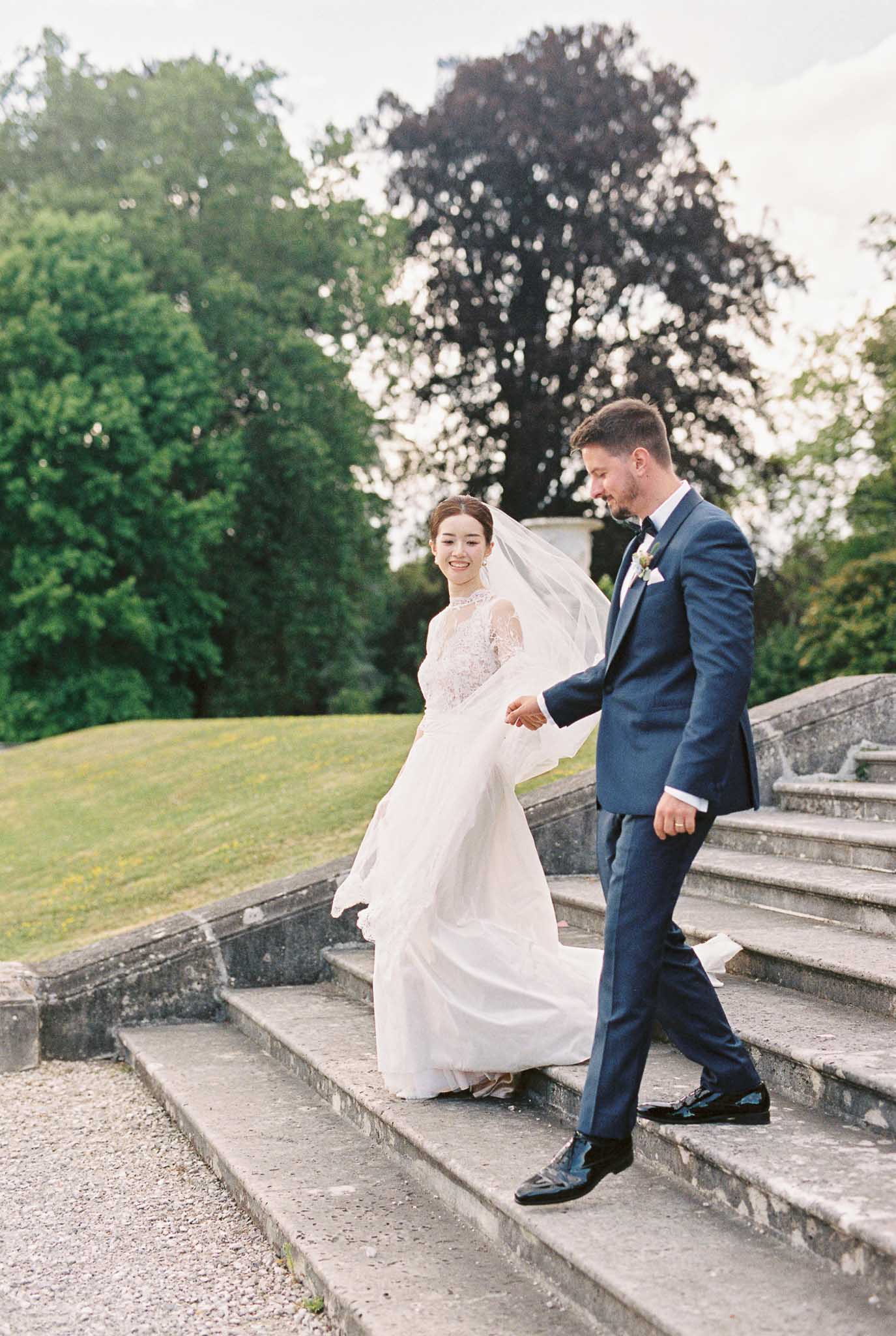 A couple portrait taken outdoors on wide stone steps leading down to a gravel path, set within formal estate grounds. The bride wears an ivory gown with a lace bodice and long sleeves, paired with a flowing cathedral-length veil caught in the wind, while the groom wears a navy blue suit with a black bow tie and a small floral boutonniere. The groom holds the bride's hand as they descend the steps together, both smiling. A classical stone urn is visible on the upper terrace behind them. The shot is a full-length portrait taken at a slight distance, capturing movement in both the skirt and veil.