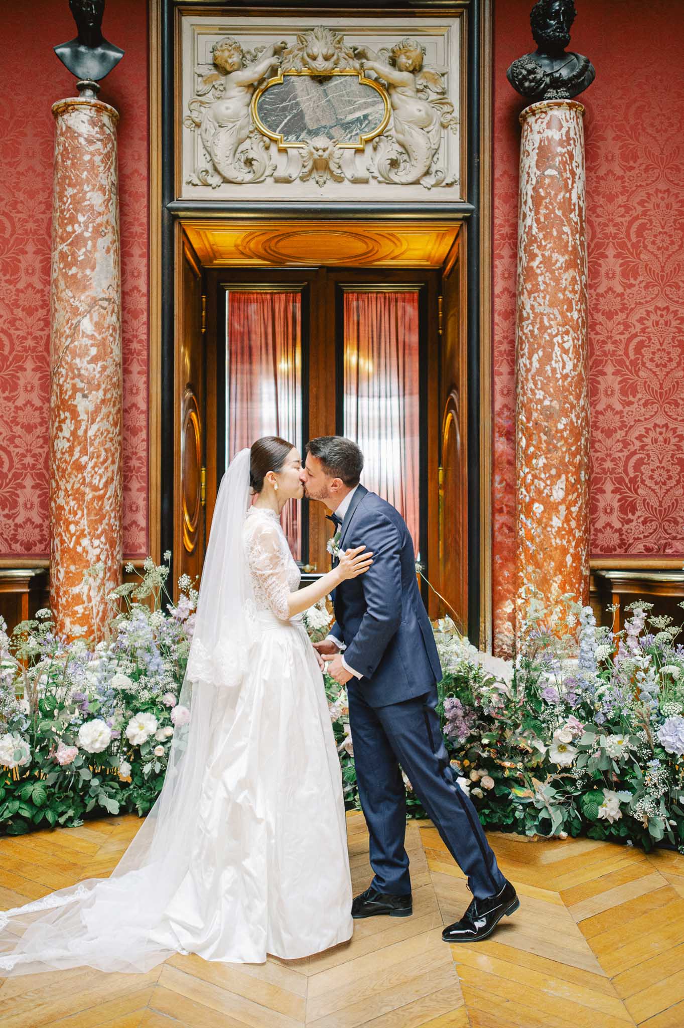 A couple shares their first kiss during an indoor ceremony in a grand classical ballroom or palace interior. The bride wears a white ball gown with lace three-quarter sleeves, a full skirt with a long train, and a cathedral-length veil, with her hair pulled back; the groom wears a navy blue suit with a bow tie and a white boutonniere. They are positioned in front of an ornate set of double wooden doors flanked by two tall veined red-and-white marble columns, with deep red damask wallcovering on the walls, gold baroque plasterwork above the doors, and dark bronze bust sculptures at the upper corners. A low garden-style floral installation runs across the base of the scene, composed of white garden roses, soft lavender and blush blooms, Queen Anne's lace, and lush greenery. The herringbone parquet wood floor is visible in the foreground. The composition is a full-length portrait shot taken at eye level.