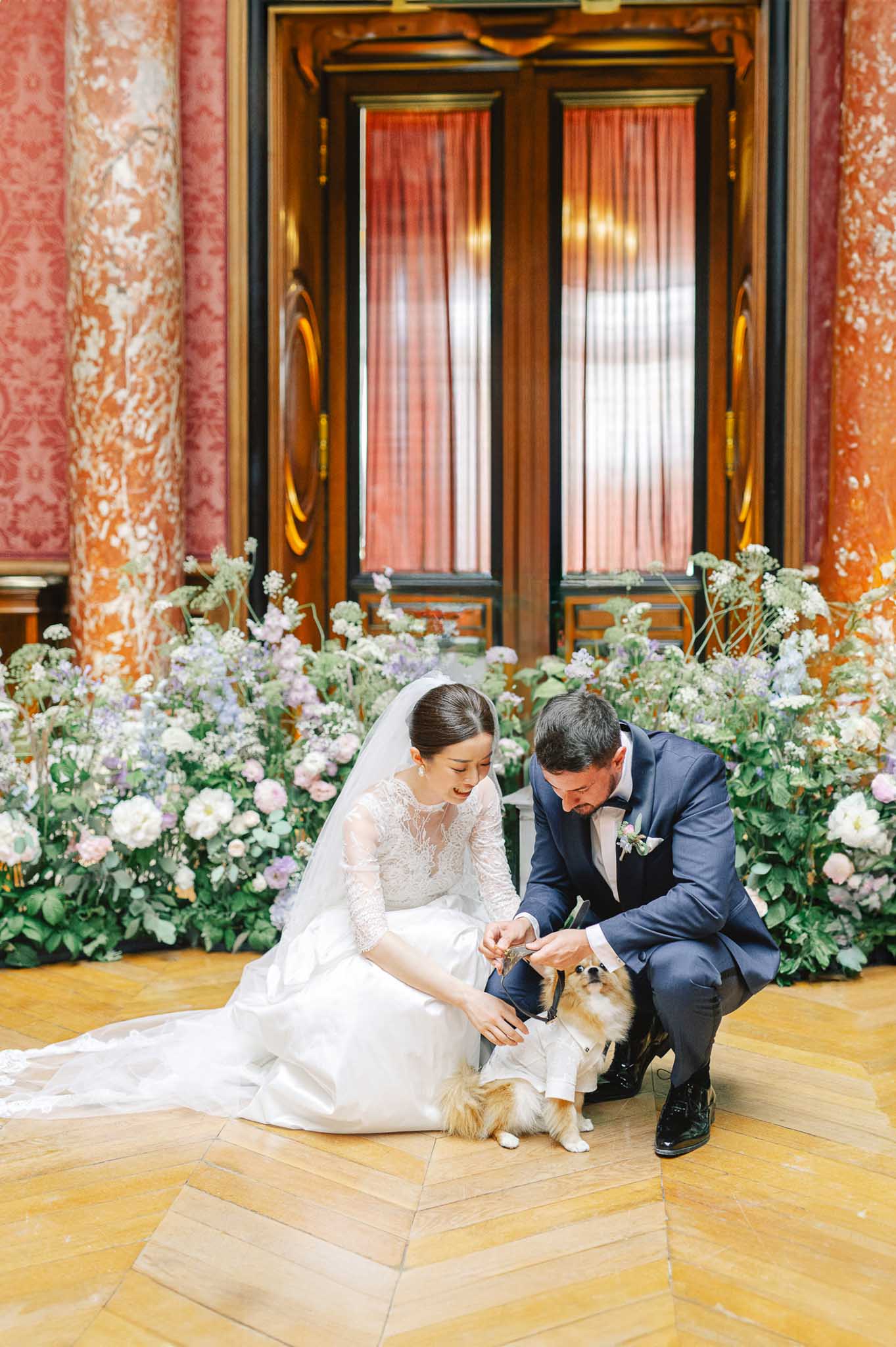 A couple portrait taken indoors in a grand hall featuring tall terracotta-toned marble columns, ornate wood-panelled double doors with coral-red curtains, and herringbone parquet flooring. The bride is seated on the floor wearing a long-sleeve lace illusion wedding gown with a full skirt and cathedral-length veil, while the groom crouches beside her in a navy blue suit with a black bow tie and a floral buttonhole; both are attending to a small Pomeranian dressed in a miniature white tuxedo outfit. Behind them, a low floral installation runs along the base of the doors and columns, featuring white peonies, soft lavender blooms, dusty mauve dahlias, white Queen Anne's lace, and abundant greenery in a garden-style arrangement. The composition is a mid-range portrait shot with the floral backdrop filling the full width of the frame.