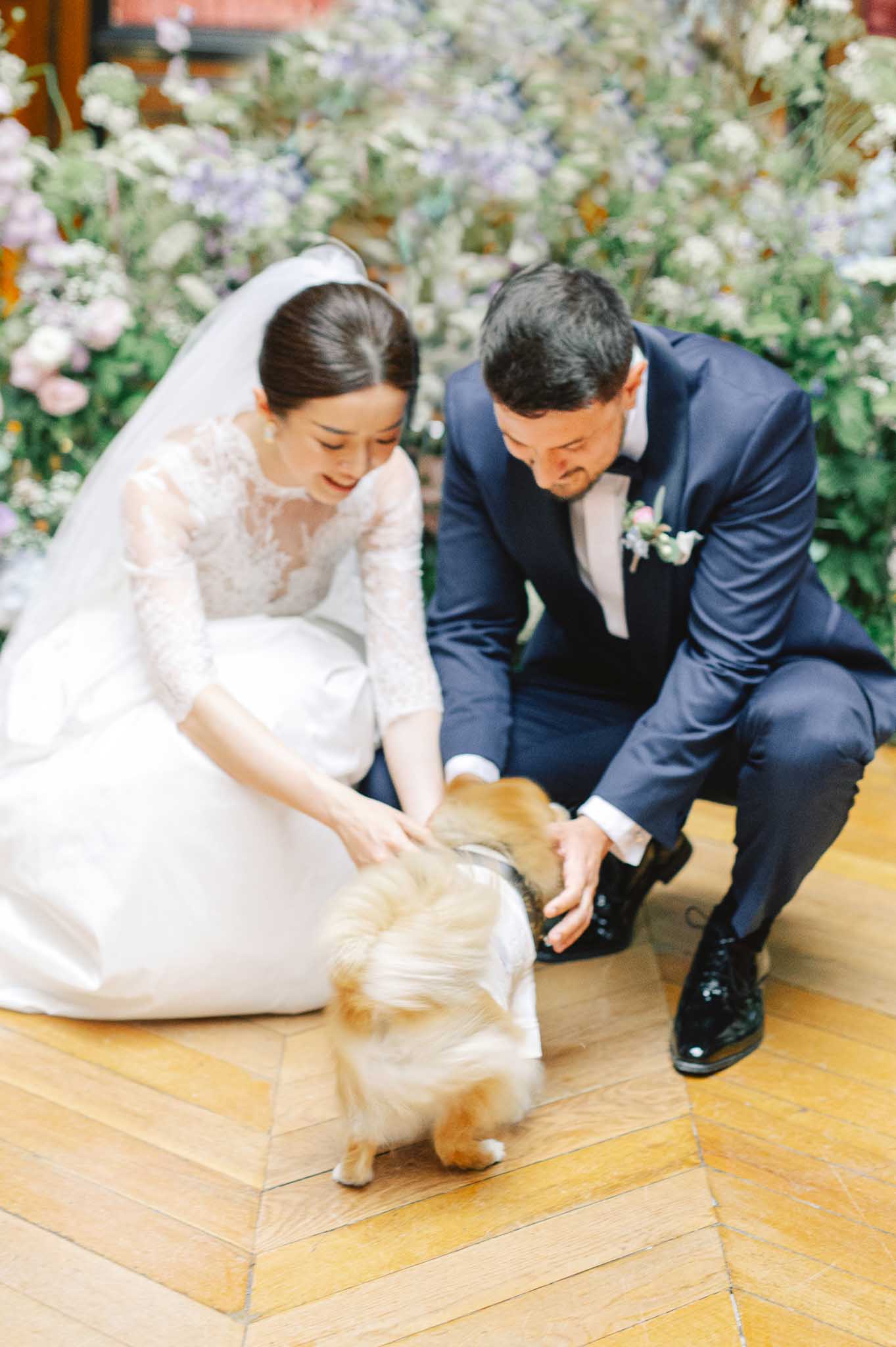 Bride and groom crouch to pet small dog before lavender and cream floral installation indoors