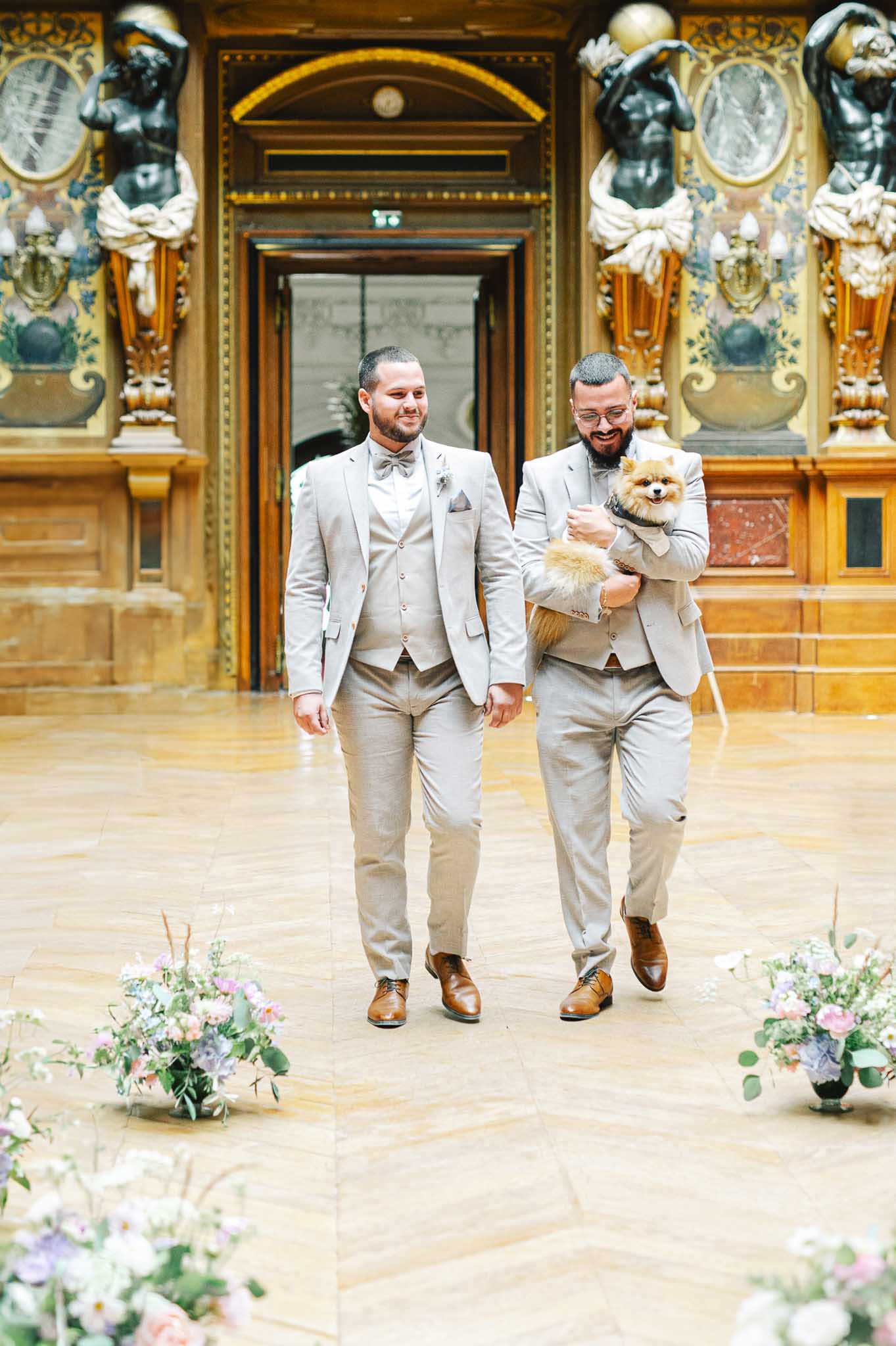 Two grooms in matching beige suits walking down aisle with Pomeranian in ornate classical hall