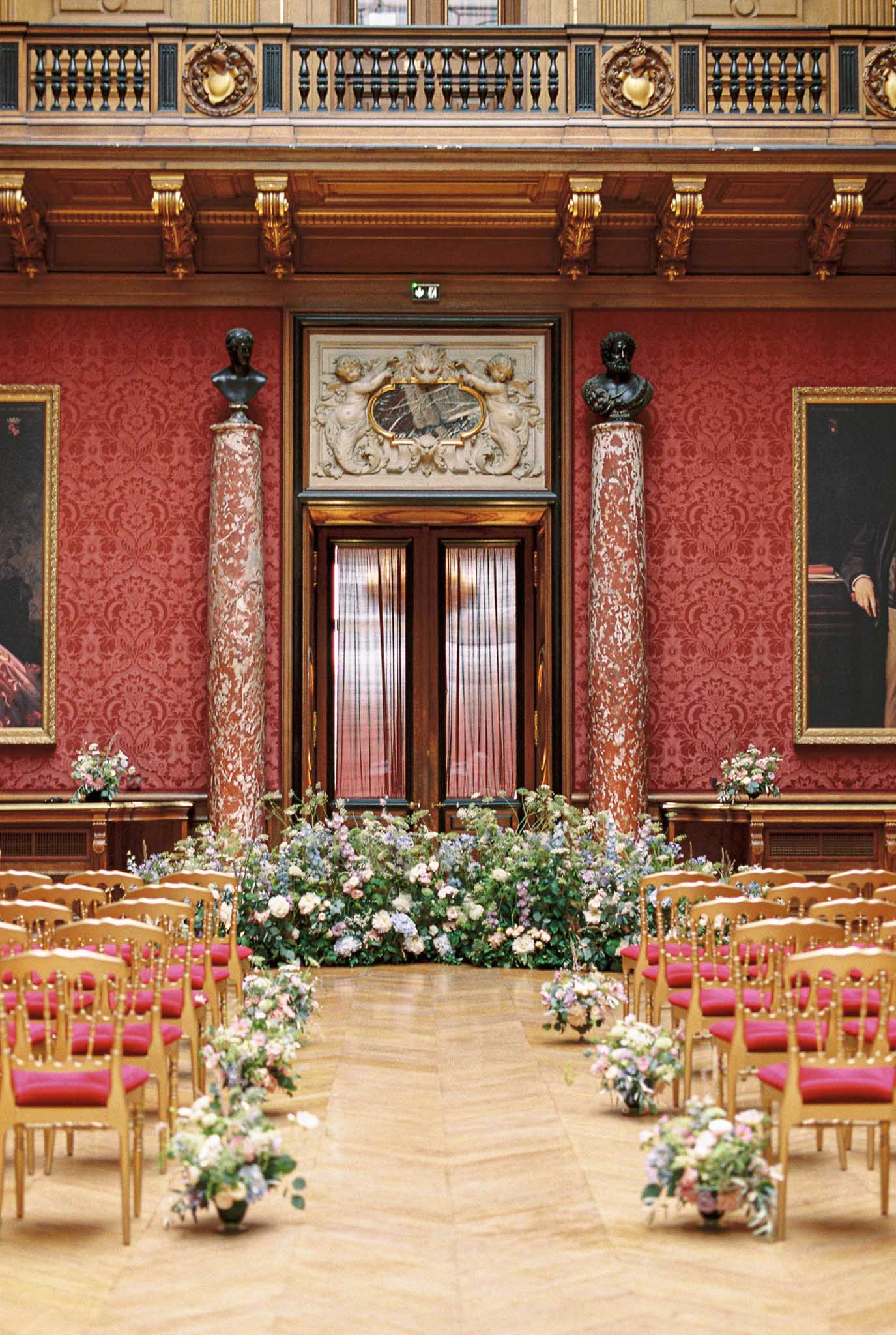 A wedding ceremony setup photographed from the aisle looking toward the altar in a grand indoor ballroom or state room with deep red damask-covered walls, warm wood paneling, and gold detailing. The space features two large variegated marble columns topped with dark bronze busts, large oil portrait paintings flanking the walls, an ornate carved stone relief above the central double doors, and a mezzanine-level balustrade with gilded accents above. Gold-framed chairs with red upholstered seats are arranged in rows on either side of a herringbone parquet aisle. Floral arrangements in blush pink, soft lavender, dusty blue, and white — appearing to include garden roses, hydrangeas, and trailing greenery — line the aisle at floor level and form a large ground-level floral installation framing the altar space in front of the doors. The overall decor palette pairs the room's existing deep red and gold architecture with soft, pastel romantic florals. Wide shot, no people present. Potential venue feature image.