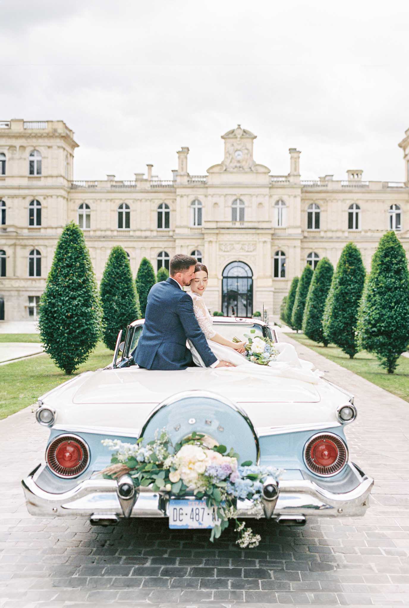 Bride and groom seated on vintage convertible with blue hydrangea bouquet in front of French chateau