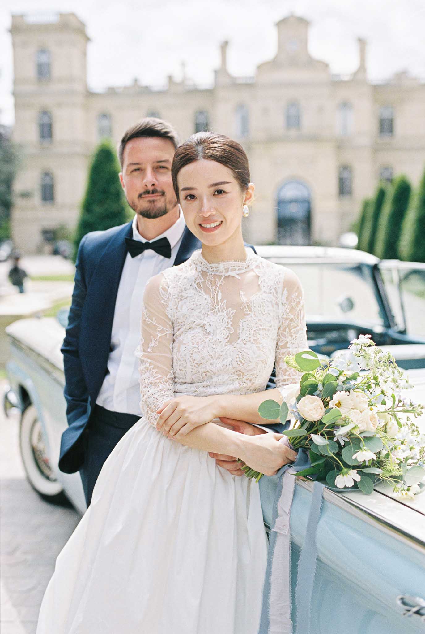 A couple portrait taken outdoors in front of a grand classical château facade with formal topiary hedges. The bride wears a two-piece ivory gown with a long-sleeved sheer lace top featuring floral appliqué and a soft tulle skirt, accessorized with pearl drop earrings and her hair pulled back in a low bun. She holds a loose, garden-style bouquet of cream garden roses, white wildflowers, and eucalyptus tied with a dusty blue silk ribbon, resting it on the door of a vintage pale blue convertible car. The groom stands behind her wearing a navy tuxedo jacket, white dress shirt, and black bow tie. The composition is a mid-length couple portrait with the château softly blurred in the background, giving the venue architectural detail context. The overall styling palette is ivory, navy, and dusty blue with a classic, refined aesthetic. Potential venue feature image.