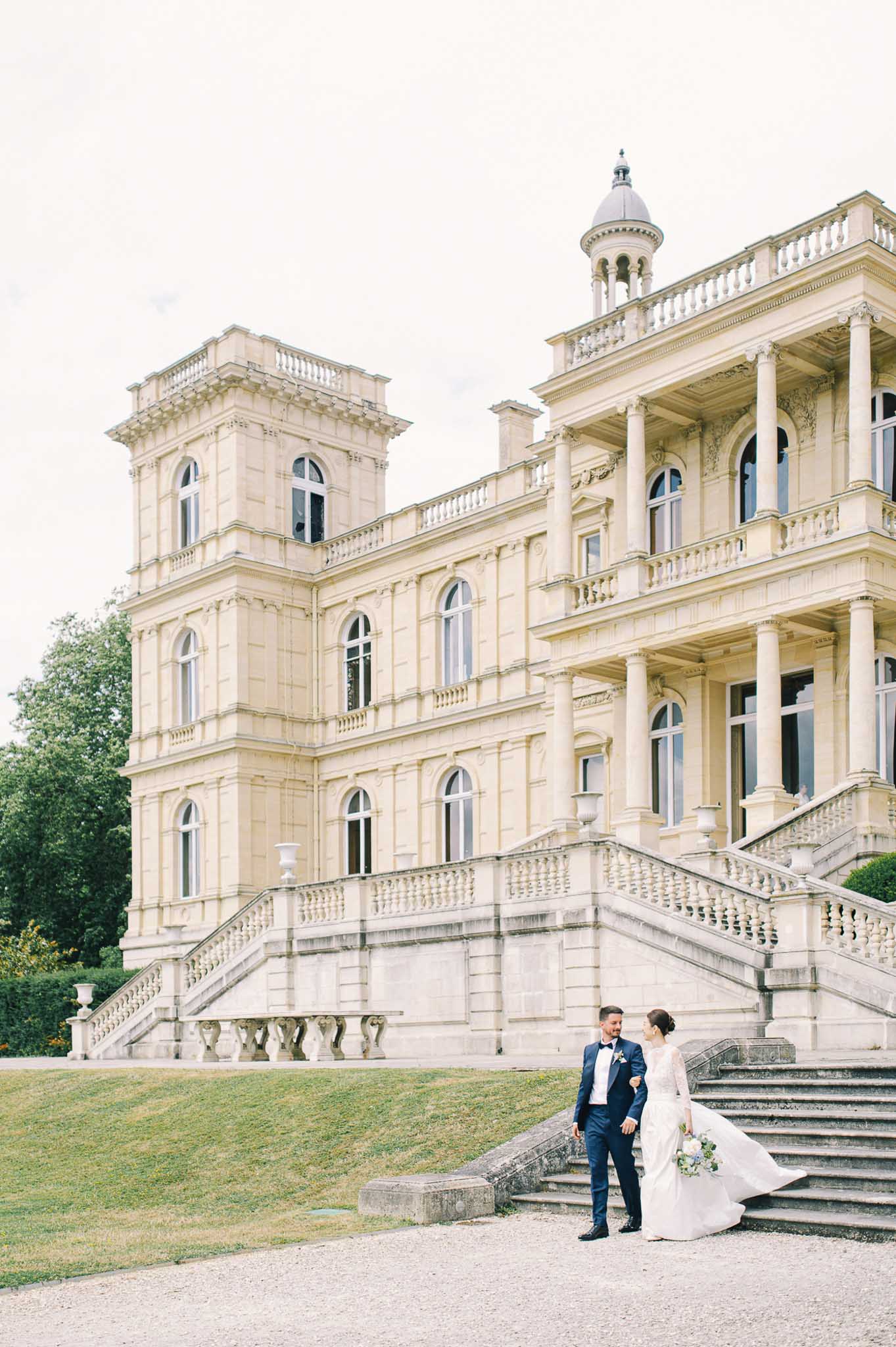 Couple walking on gravel before grand chateau with double staircases bride carrying blue and white bouquet