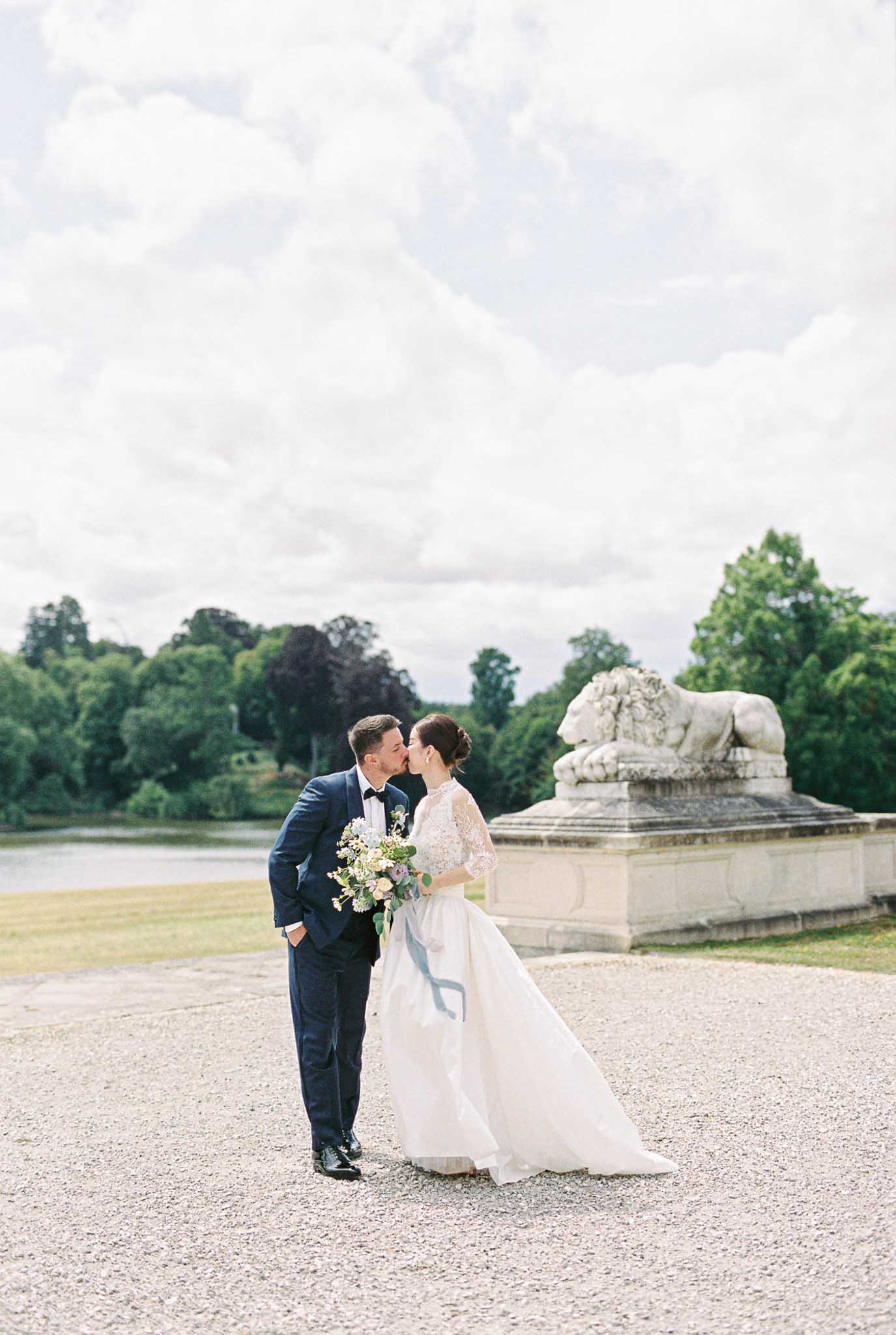 A couple portrait taken outdoors on a gravel path, with the bride and groom sharing a kiss. The groom wears a navy blue suit with a black bow tie and boutonniere, while the bride wears a white ballgown with a lace long-sleeved bodice and a full tulle skirt with a trailing hem. She carries a loose, garden-style bouquet of blush and ivory blooms with eucalyptus and greenery, tied with a long dusty blue silk ribbon. A large stone lion sculpture on a raised pedestal is positioned to the right of the couple, and a reflective body of water is visible in the background. The shot is a full-length portrait framed in a classic, film-photography style with soft, airy tones consistent with film stock.
