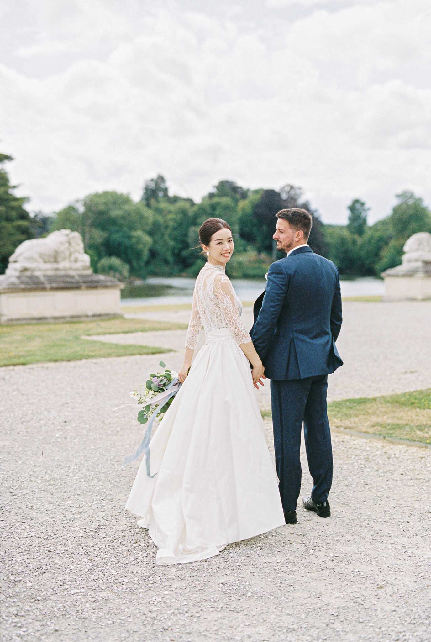 Couple on gravel path with bride in lace-sleeve ballgown holding eucalyptus bouquet with blue ribbons