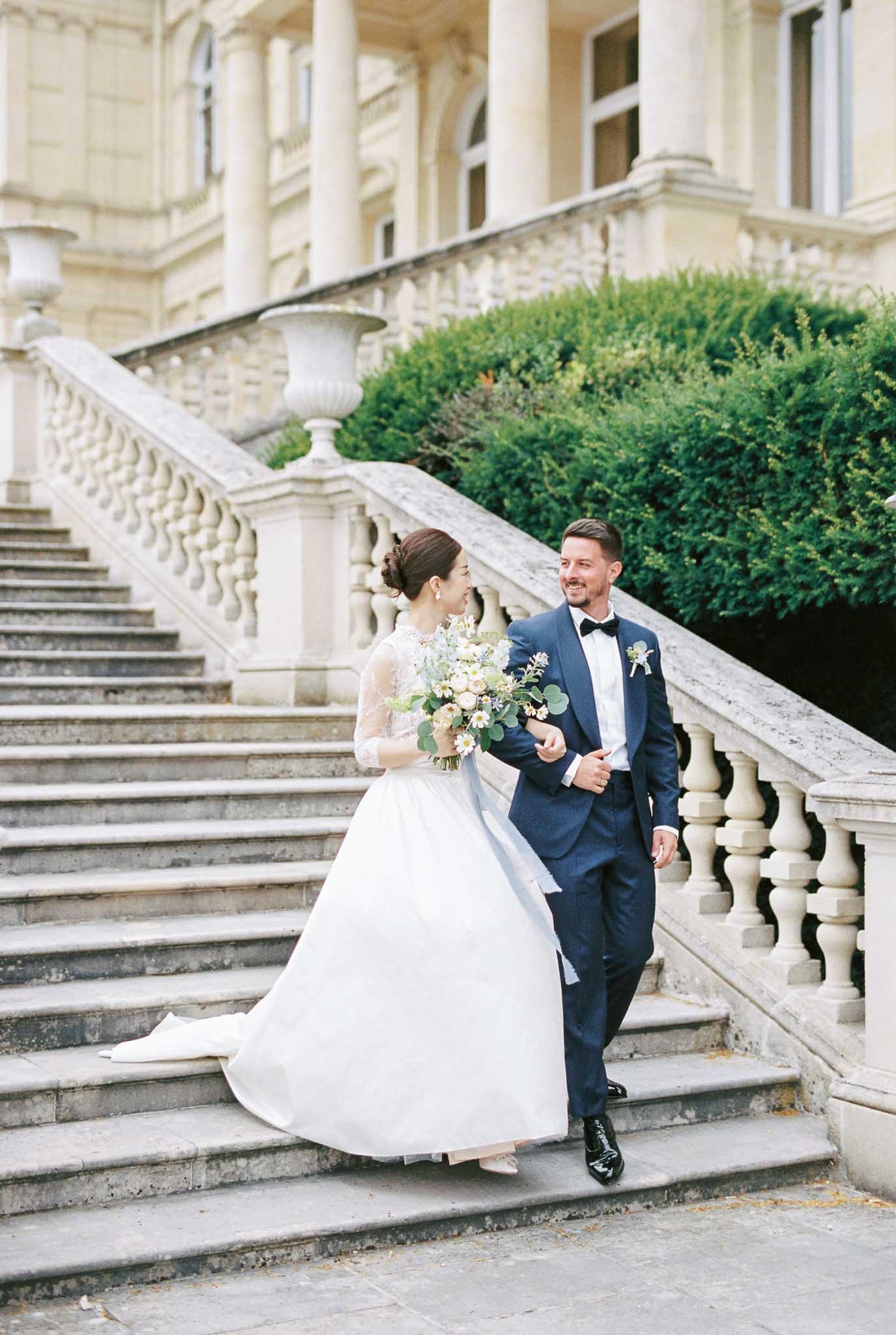 Couple walking down chateau stone staircase bride with daisy bouquet and blue ribbon groom in navy suit