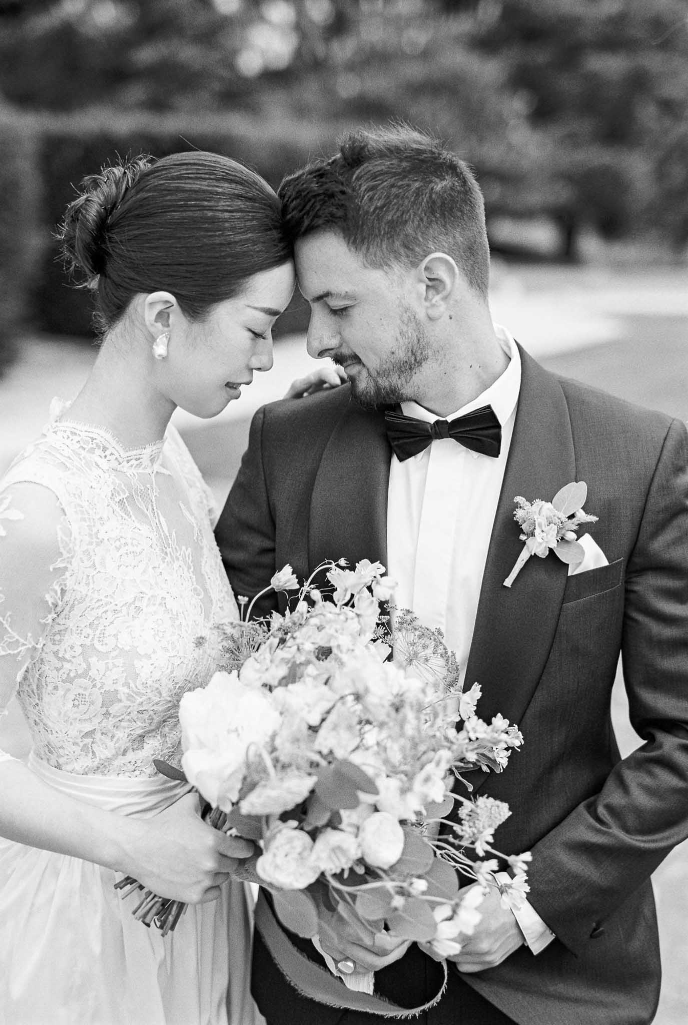 Black and white intimate close-up foreheads touching bride in lace-sleeve gown with bouquet groom in suit with pocket square