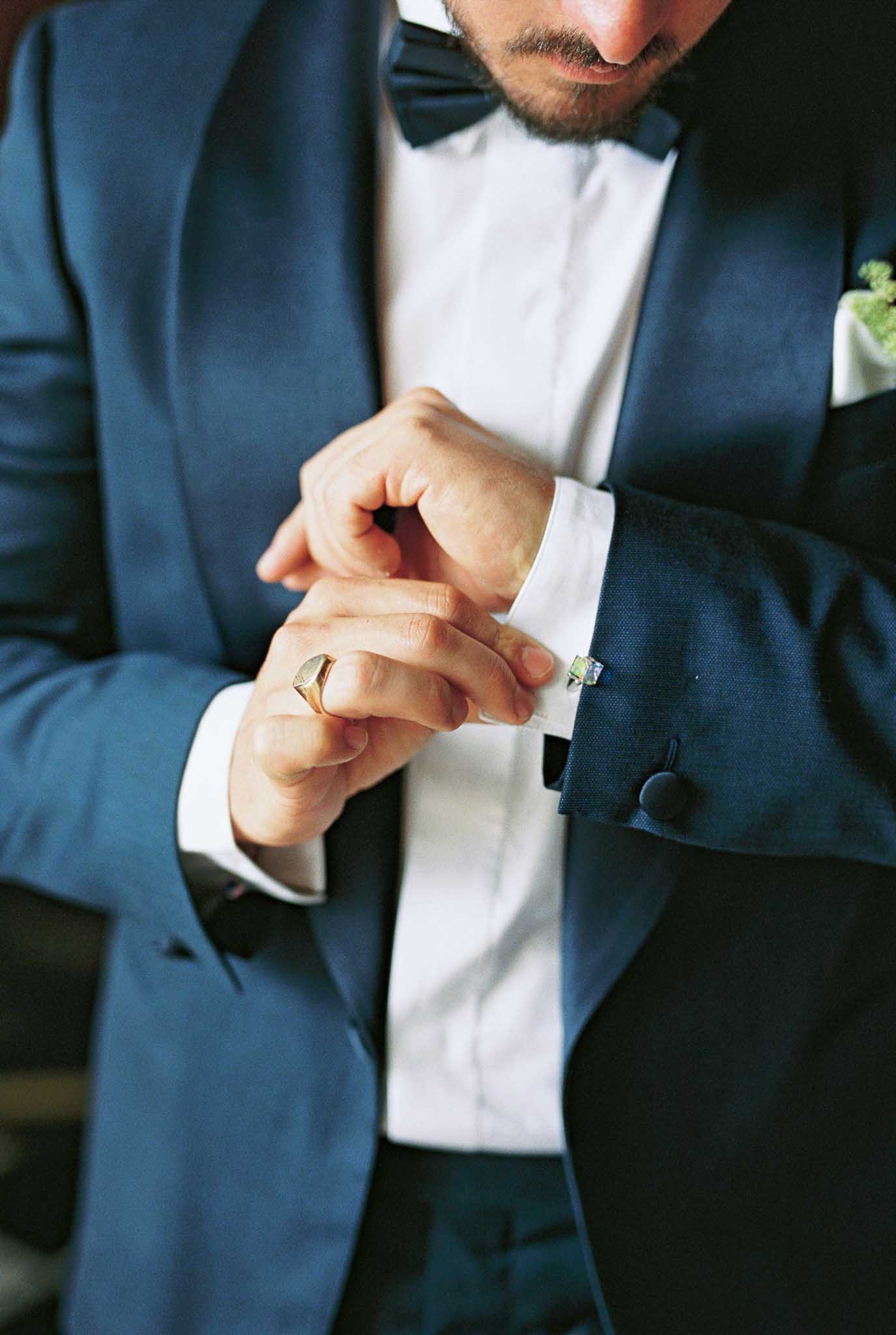 Groom fastening blue stone cufflinks wearing navy suit with bow tie and green boutonniere