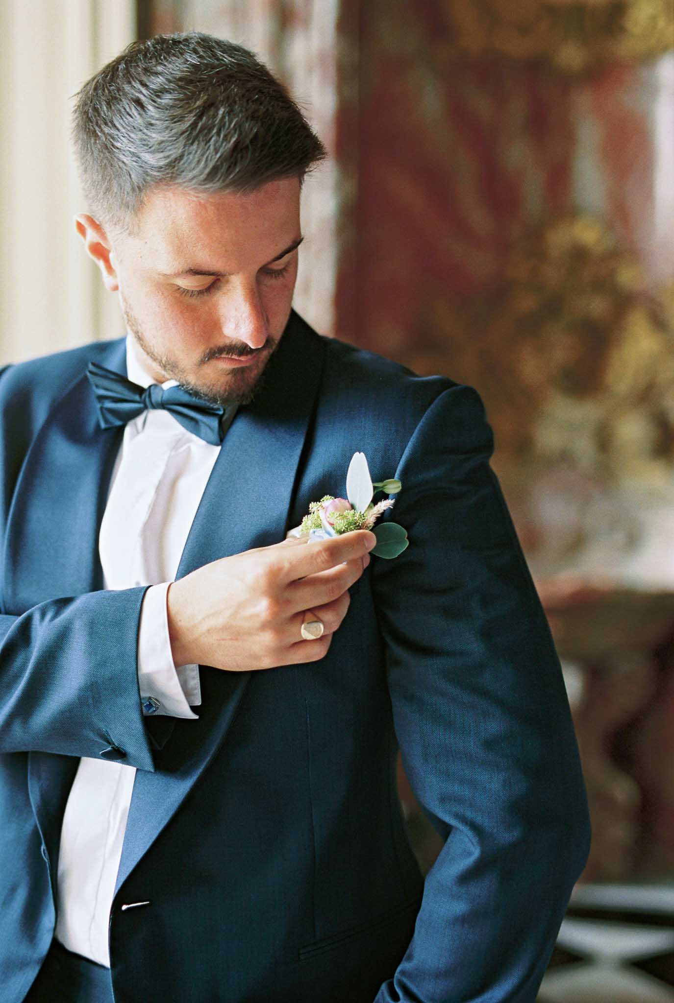 A groom is photographed indoors during the getting-ready phase, adjusting a boutonniere on the lapel of his navy blue tuxedo jacket. He wears a matching navy bow tie, a white dress shirt with French cuffs and blue cufflinks, and a signet ring on his right hand. The boutonniere features a small white bloom, a blush or mauve accent flower, green foliage, and what appears to be a sprig of dried grass or pampas. The background shows ornate wall paneling with warm red and gold tones, suggesting a classical interior such as a château or historic venue. The shot is a close-up portrait with shallow depth of field, keeping focus on the groom's face and boutonniere while the background is softly blurred.