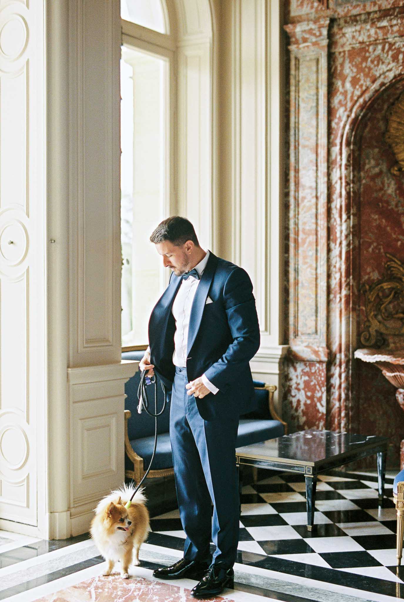 Groom in navy tuxedo with Pomeranian on leash in marble-floored chateau salon with gilded fireplace