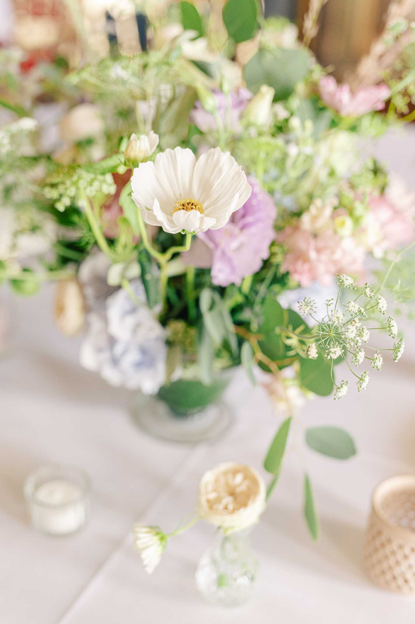 Glass vase centerpiece with white cosmos, lavender sweet peas, blue hydrangea, and eucalyptus on white linen