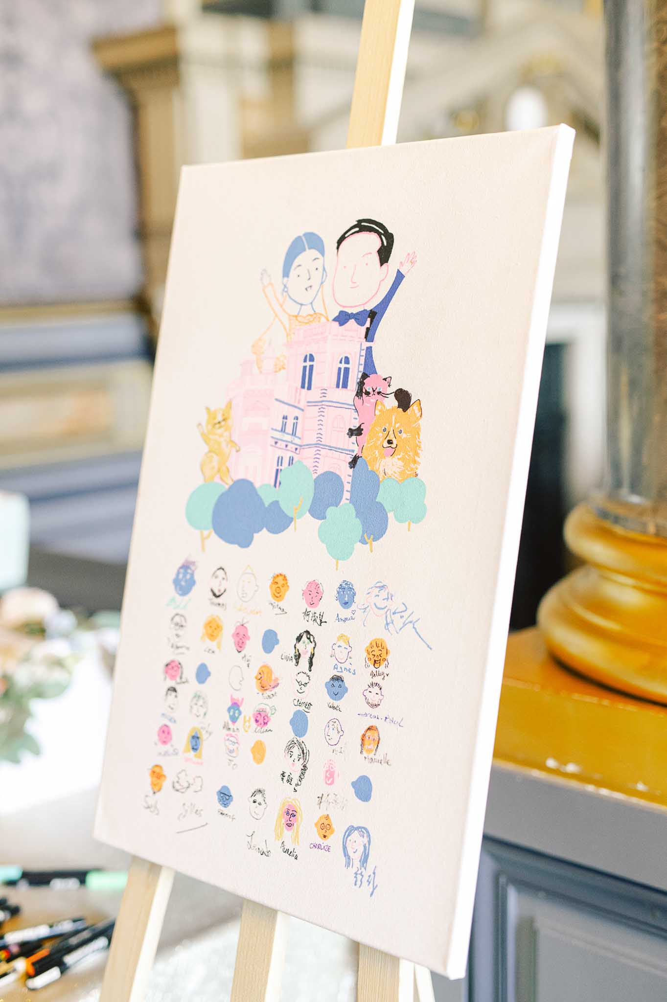Illustrated wedding guest book canvas on wooden easel with hand-drawn couple, chateau, and guest portrait signatures