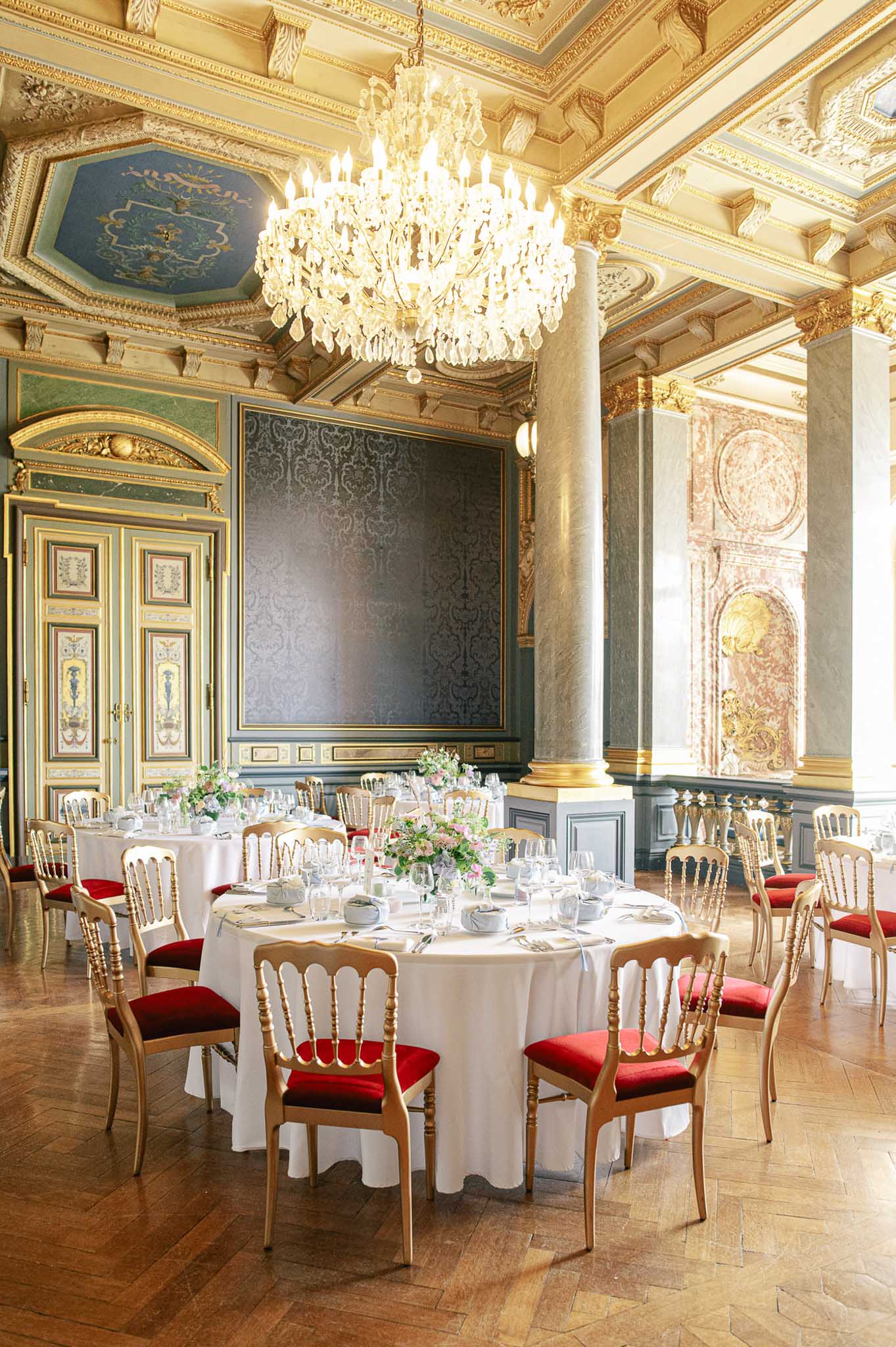 A wide interior shot of a grand ballroom set up for a wedding reception, with no guests present. Two round tables are dressed in white floor-length linen and set with glassware, plates, and folded napkins, centered with low floral arrangements featuring soft pink blooms and green foliage. The chairs are gold Napoleon-style with deep red velvet seat cushions. The room features ornate gold-detailed ceiling moldings, a painted blue ceiling panel, a large crystal chandelier, damask-paneled walls in muted blue-grey with gold framing, marble-effect columns with gold bases, and herringbone parquet flooring. The overall décor style is classic French palatial, with the venue's architectural detailing serving as the dominant design backdrop. Potential venue feature image.