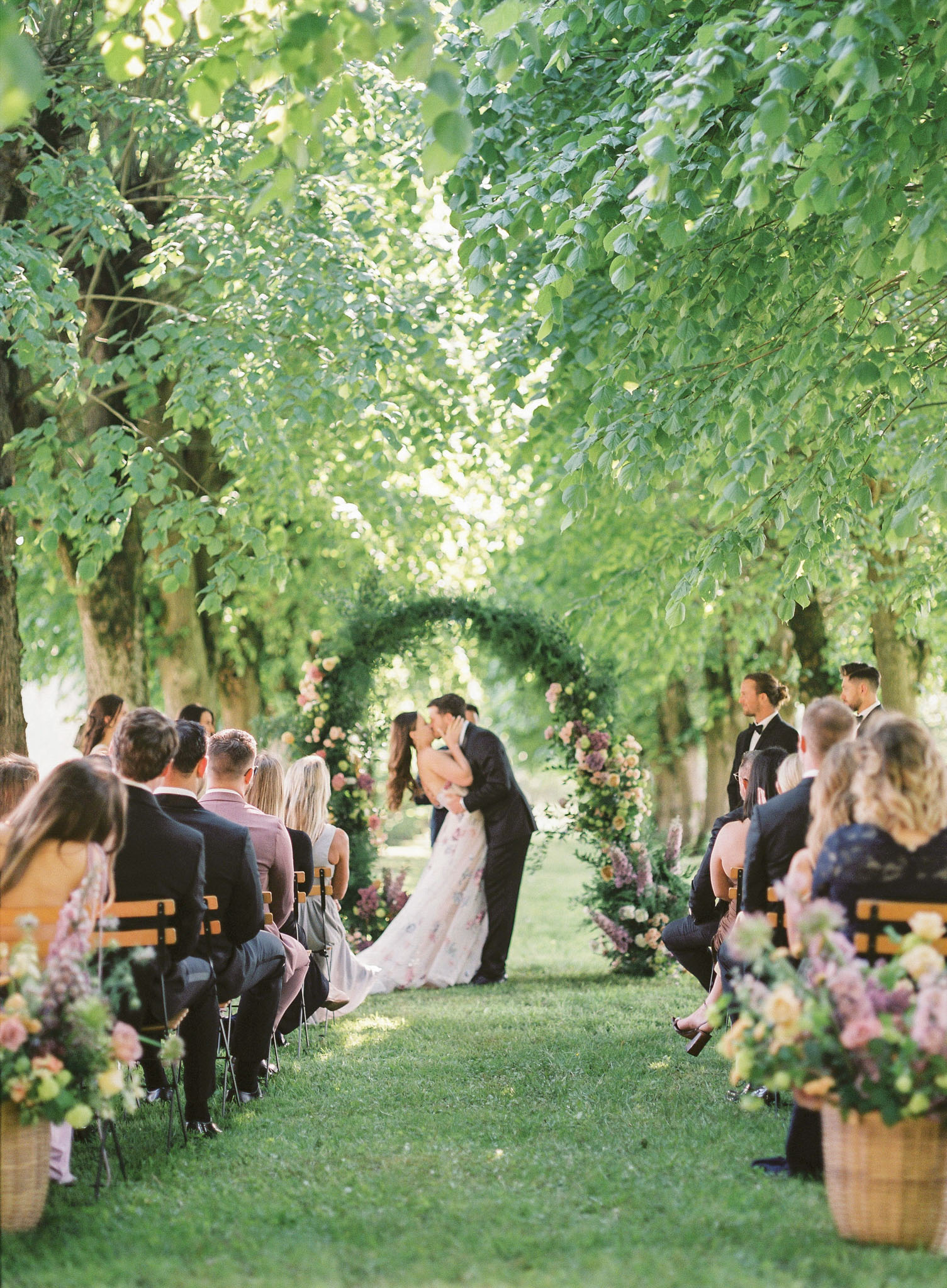 Couple shares first kiss under a circular floral arch during an outdoor garden ceremony with seated guests