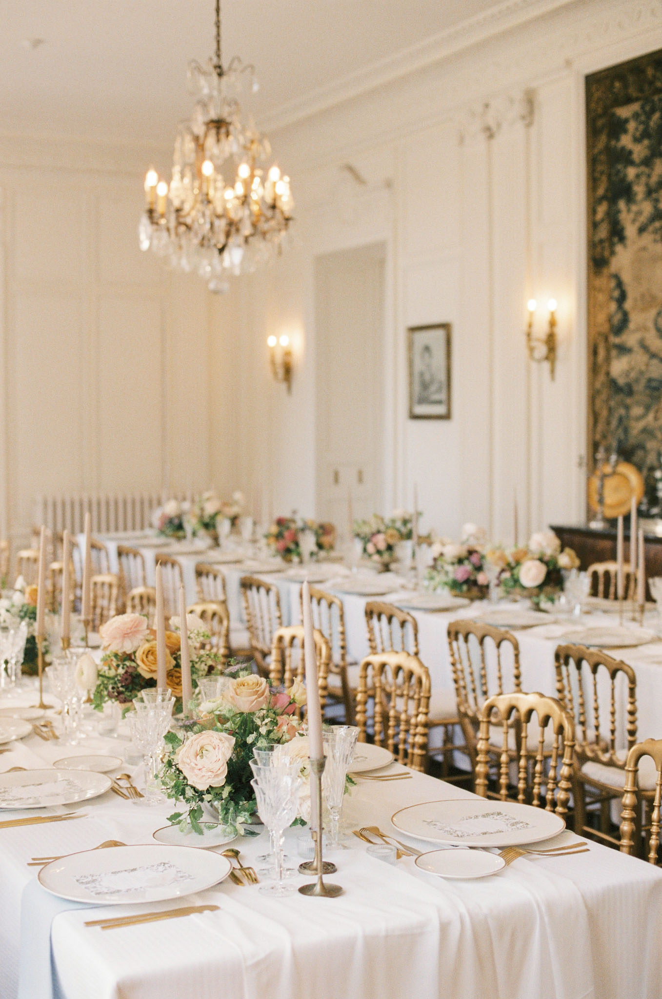 A reception tablescape detail shot taken inside a formal French château ballroom with white panelled walls, gold wall sconces, a crystal chandelier, and a large tapestry visible on the far wall. Multiple long banquet tables are dressed in white linen and lined with gold Napoleon-style chairs, set for what appears to be a seated dinner for a large number of guests. Each place setting features white porcelain plates with gold-rimmed edges and handwritten calligraphy menus, gold flatware, and cut-crystal glassware. Low floral centerpieces of blush garden roses, peach and amber roses, and trailing greenery are interspersed with slim taupe taper candles in gold and silver candlestick holders. The overall decor palette is ivory, gold, and soft blush with a classic French formal styling. Potential venue feature image.