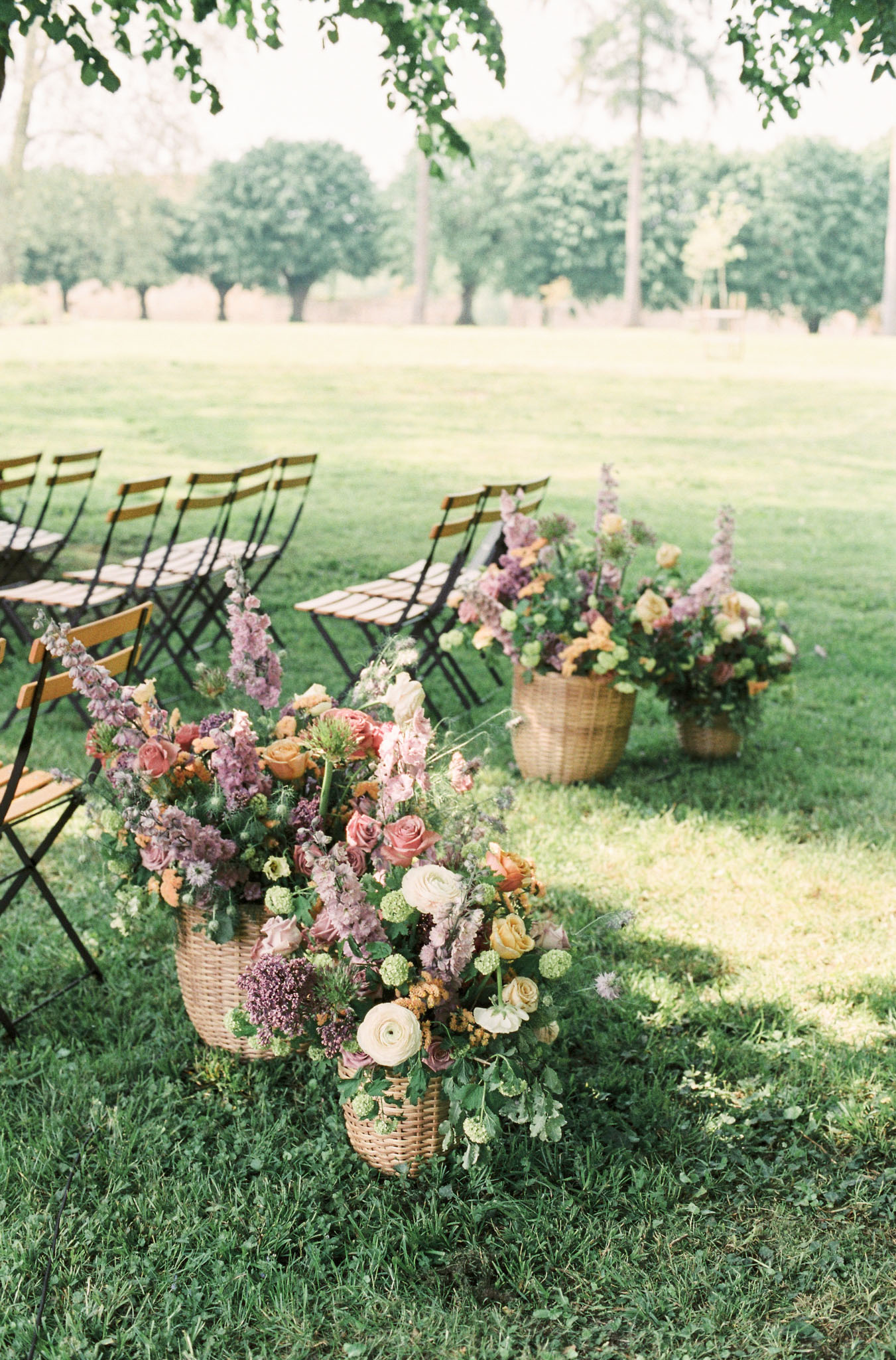 Outdoor ceremony aisle with wicker basket floral arrangements in mauve and blush tones and wooden folding chairs on lawn
