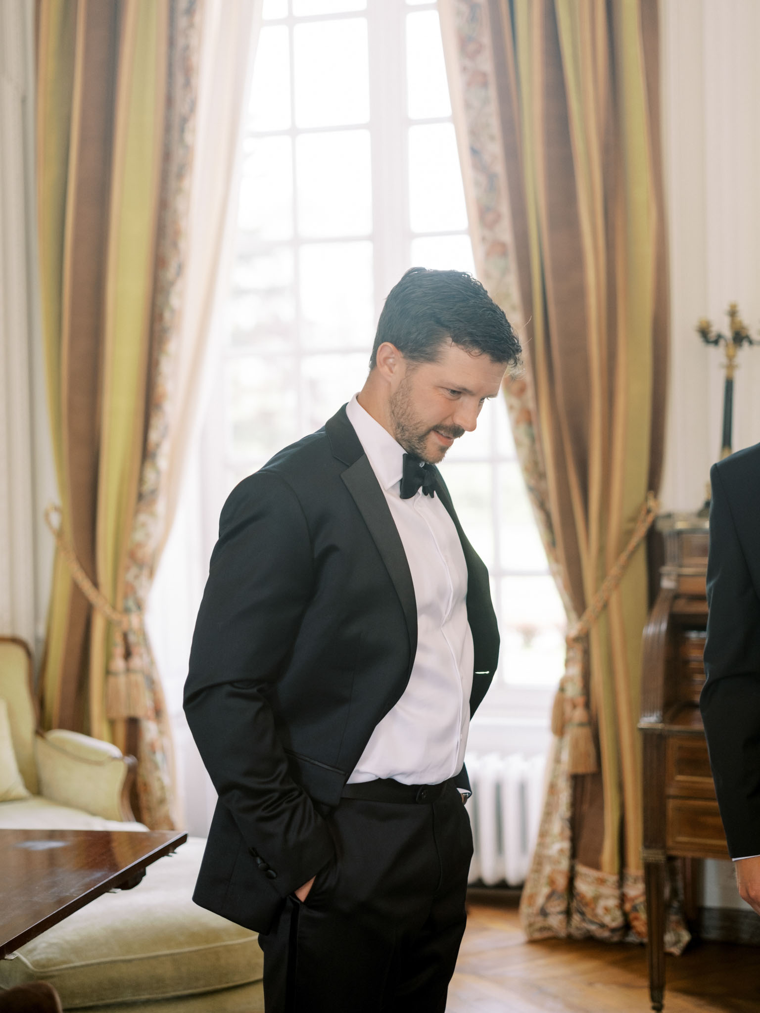 Groom in tuxedo with satin lapels standing in chateau room with gold curtains and parquet floor