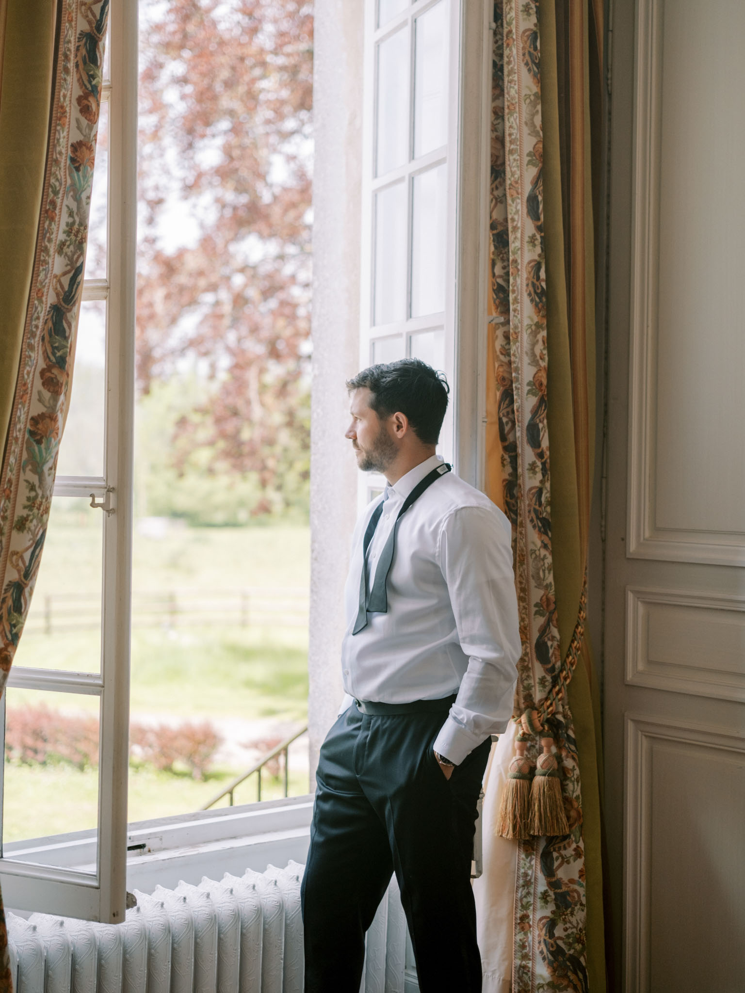 A groom is captured getting ready, standing at an open French window and gazing outside at the grounds below. He is dressed in a white dress shirt with dark navy trousers, black suspenders, and an untied dark tie hanging loosely around his collar, suggesting the shot was taken mid-preparation. The interior setting features classic French château-style details including white wood-panelled walls, a cast-iron radiator, and floor-length curtains in a warm gold and floral toile fabric with gold tassel tie-backs. The shot is a medium portrait framed in profile, with natural window light illuminating the groom against the soft-focus garden view beyond.