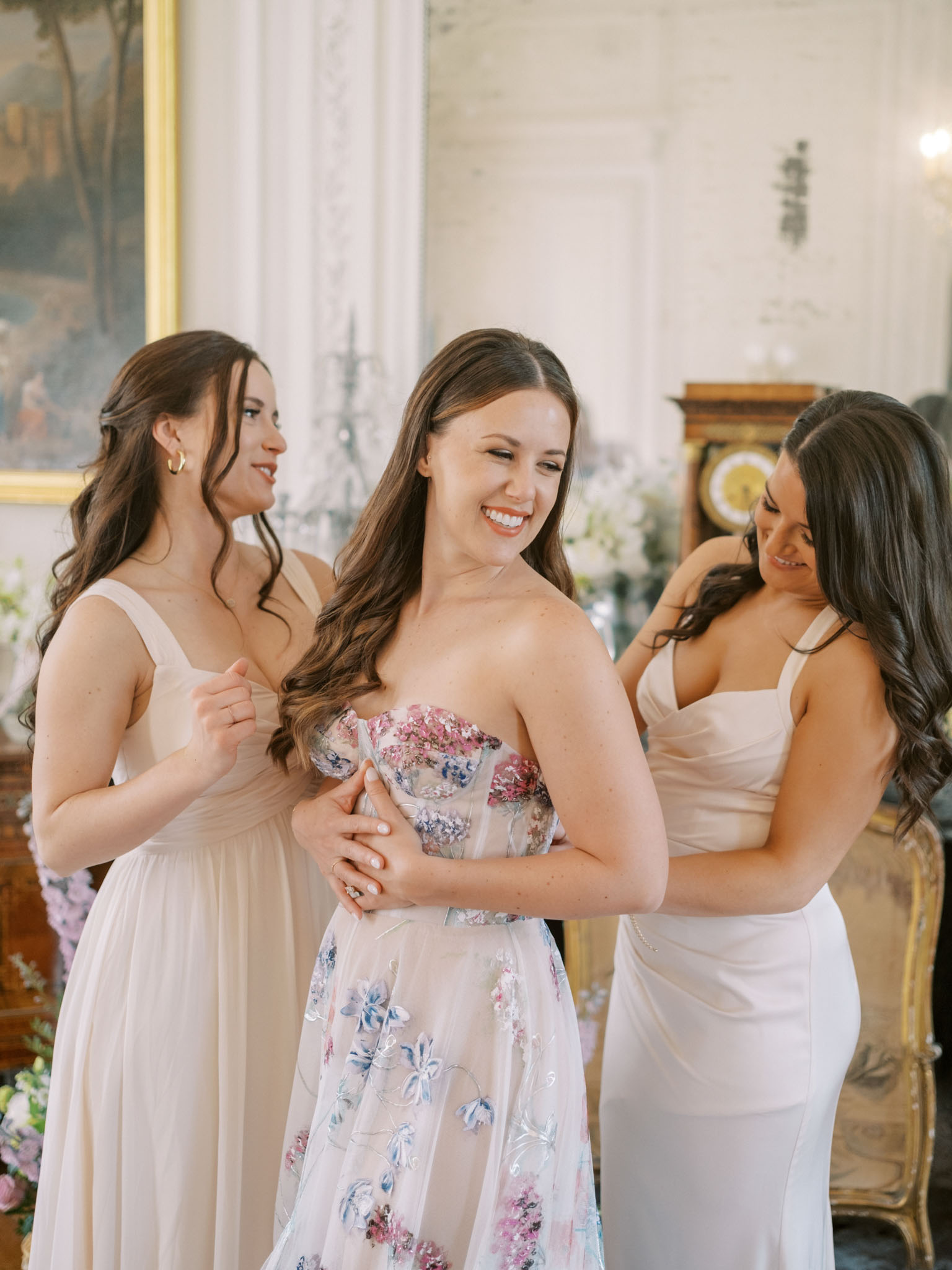 Bride in strapless floral embroidered gown smiling as two bridesmaids fasten her dress in an ornate salon