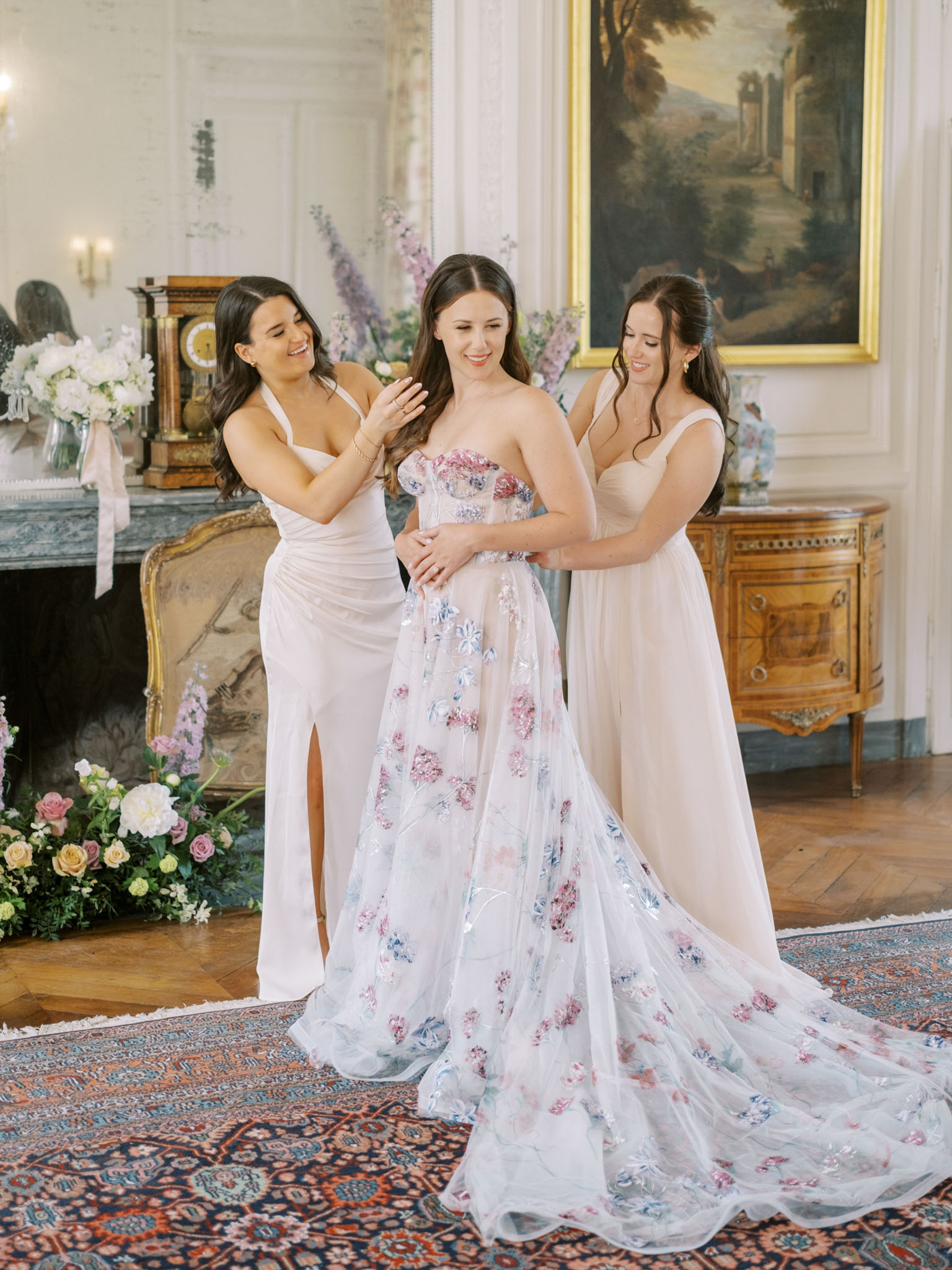 A getting-ready moment inside a French château salon, where two bridesmaids help the bride with final adjustments to her look. The bride wears a strapless A-line gown in ivory tulle with an all-over floral appliqué pattern in dusty pink, mauve, and periwinkle blue, with a full trailing skirt. The two bridesmaids wear blush pink floor-length dresses — one in a halter-neck style with a front slit, the other in a wide-strap column style. The room features classic French interior details including a carved marble fireplace, herringbone parquet flooring, an ornate patterned area rug, a gilded oil painting, and a Louis XVI-style commode. Floor-level floral arrangements of blush roses, peach roses, ivory blooms, and eucalyptus are placed near the fireplace, and tall stems of lavender-blue delphiniums are visible on the mantel alongside white floral arrangements tied with blush ribbon. The overall styling palette is soft blush, white, and muted botanicals in a classic French interior setting. Medium-distance portrait shot framing all three women.
