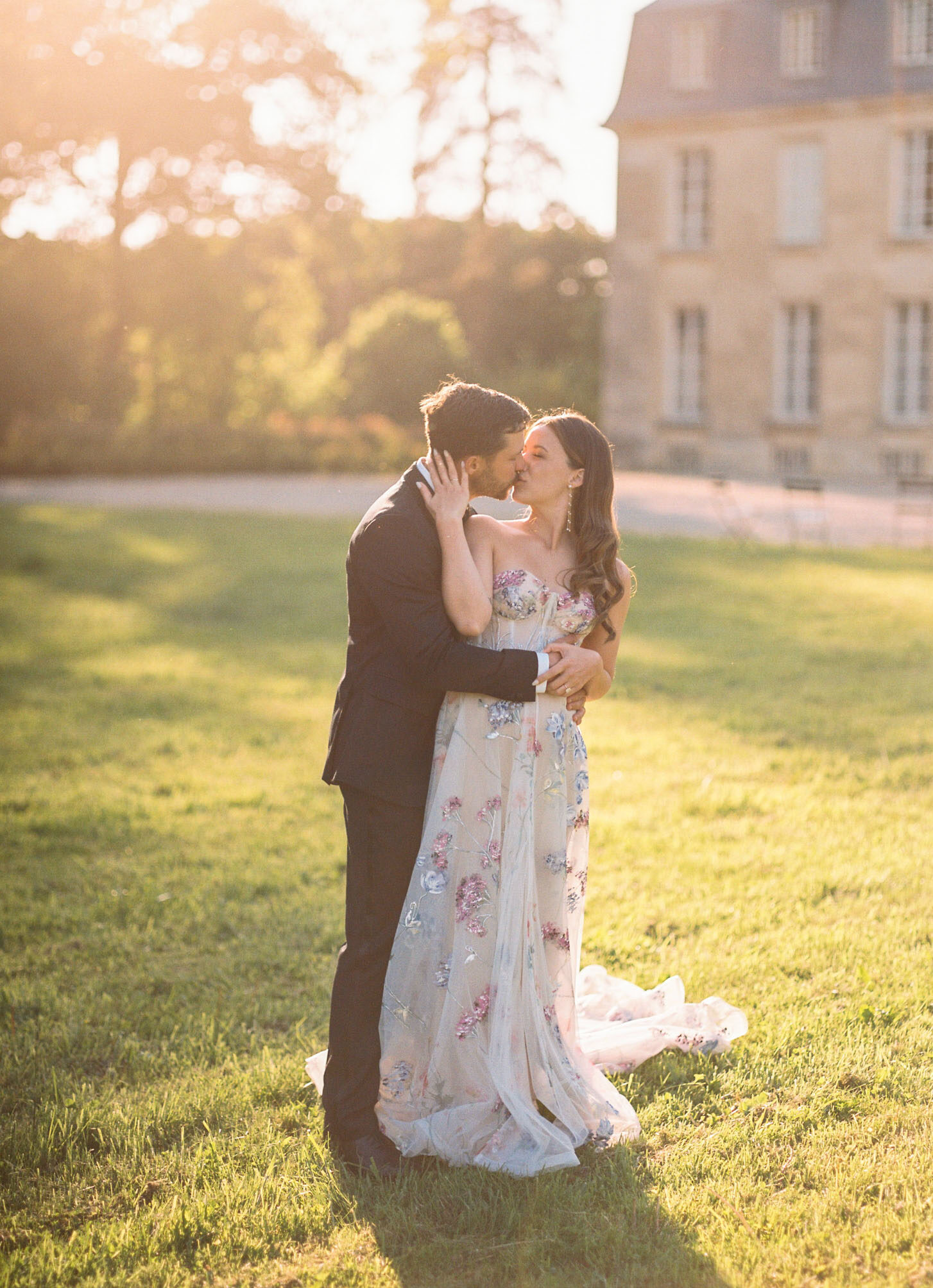 Couple kisses at golden hour with bride's floral-applique gown train spread across lawn at chateau